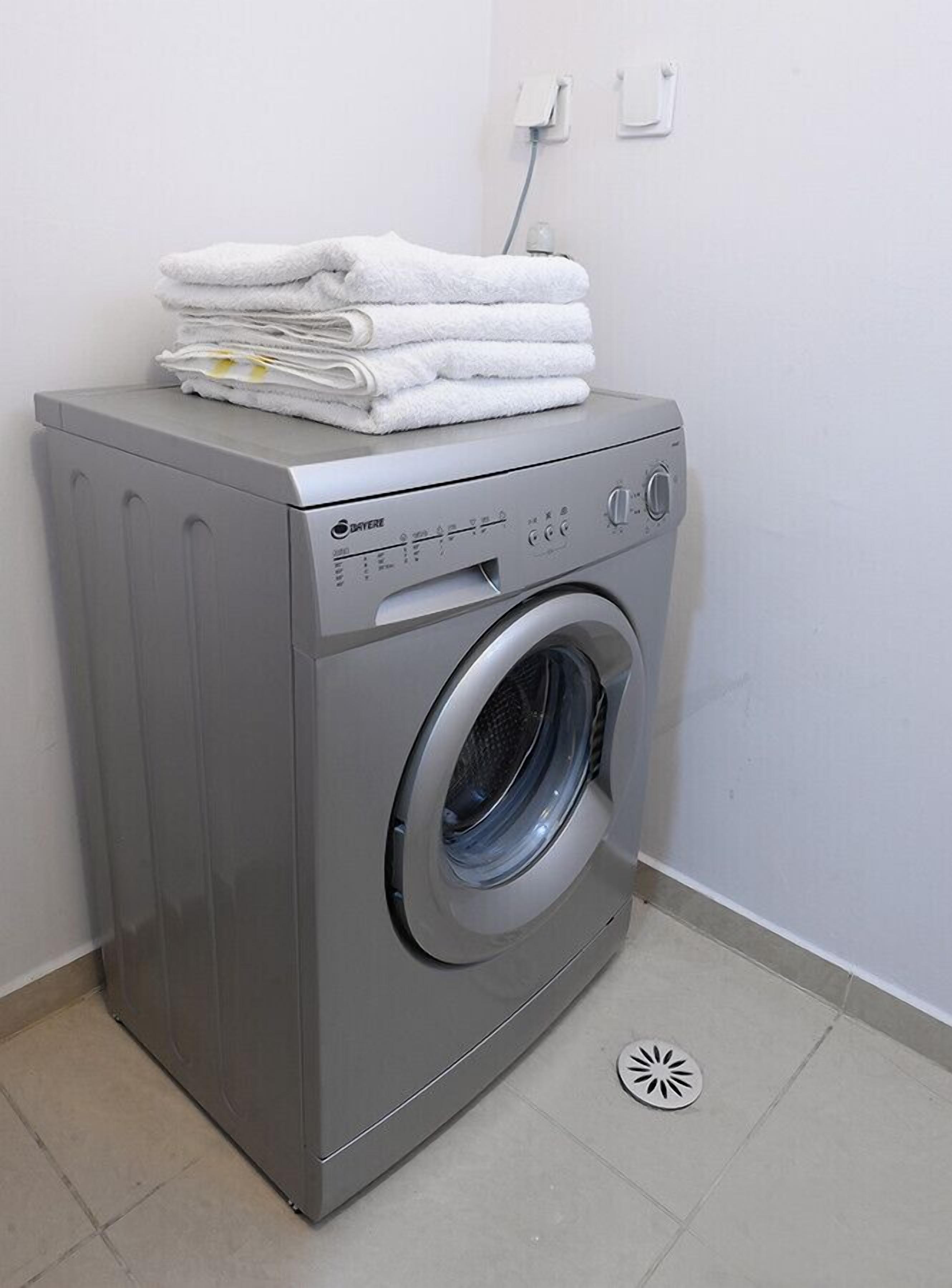A front-loading gray washing machine with a stack of white towels on top, set against a plain white wall with a power outlet.