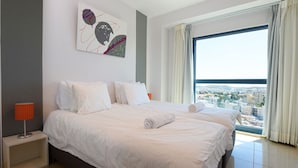 A hotel room with a large bed, a nightstand, a lamp, a painting, and a view of the city through a large window.
