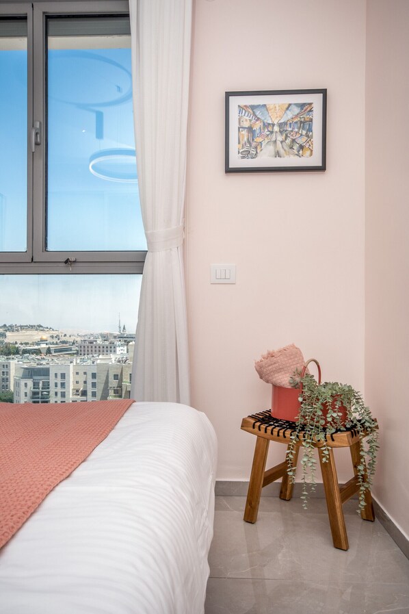 Une chambre à coucher avec un lit, une fenêtre avec des rideaux, un tableau encadré au mur et un petit tabouret en bois sur lequel est posée une plante.
