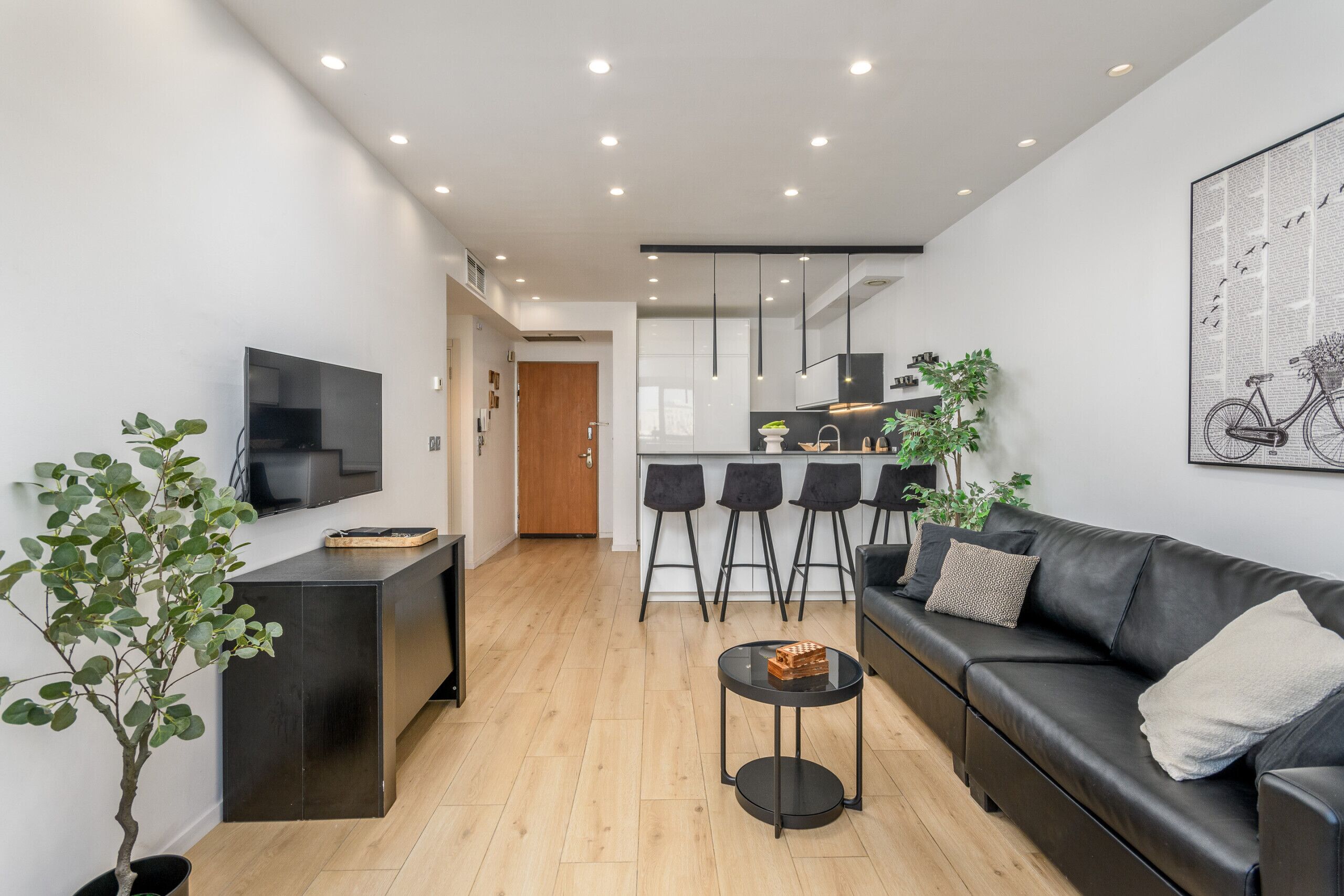 Ein modernes Wohnzimmer mit einem schwarzen Ledersofa, einem runden Couchtisch und einem an der Wand montierten Fernseher. Es gibt eine Küchenzeile mit Barhocker und einer Topfpflanze.