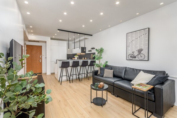 Una sala moderna con un sofá de cuero negro, una mesa de centro y una zona de comedor con taburetes de bar.