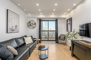 Una sala de estar moderna con un sofá de cuero negro, una mesa de centro redonda, un televisor y un balcón con vistas a edificios.