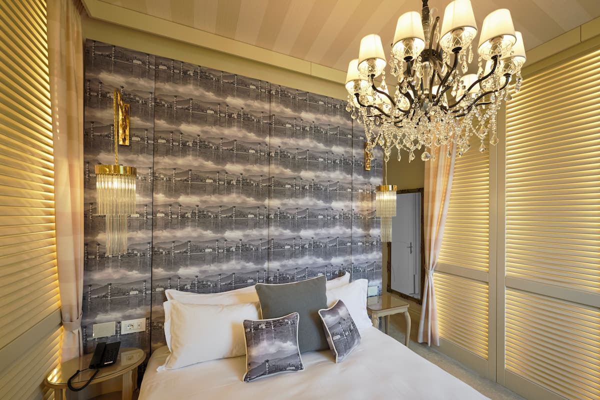 Habitación de hotel con una cama grande, una araña de cristal, un teléfono en una mesita de noche y una pared con un diseño repetido.