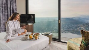 Ein Hotelzimmer mit einem großen Bett, einem Fernseher, einem Frühstücksbrett und Blick auf Stadt und Berge.