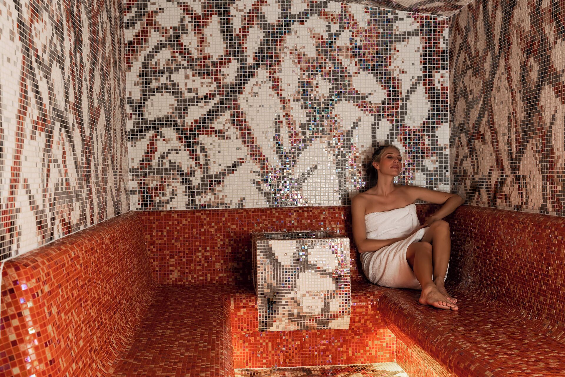 Una persona seduta in una stanza con piastrelle a mosaico, una parete decorata e una panca in piastrelle rosse.