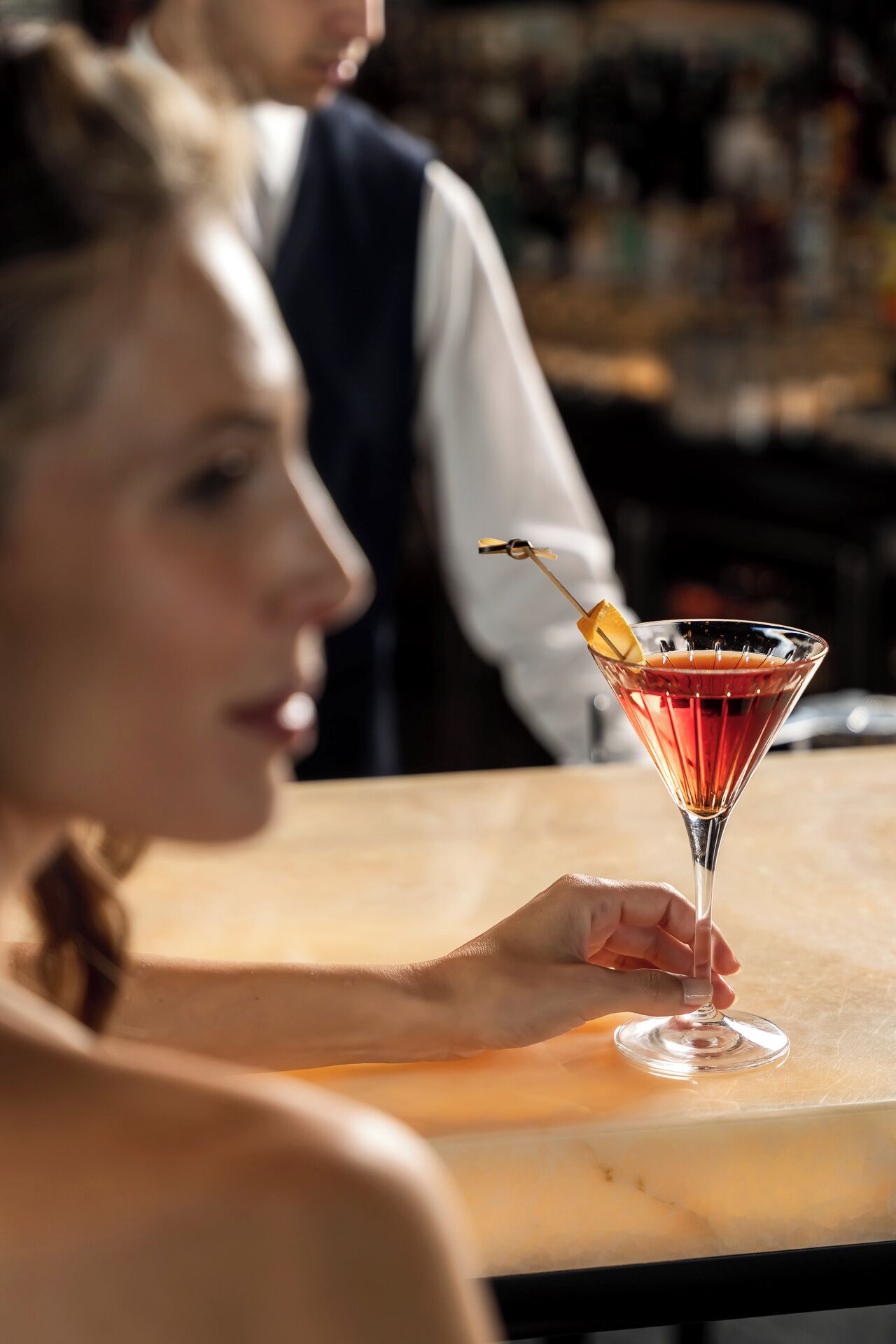 A person holding a martini glass with a lemon twist on a bar counter.