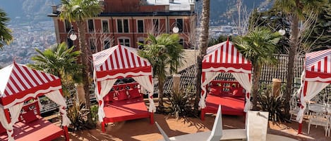 A pool area with red and white striped canopies, a clear view of a cityscape in the background, and a palm tree in the foreground.
