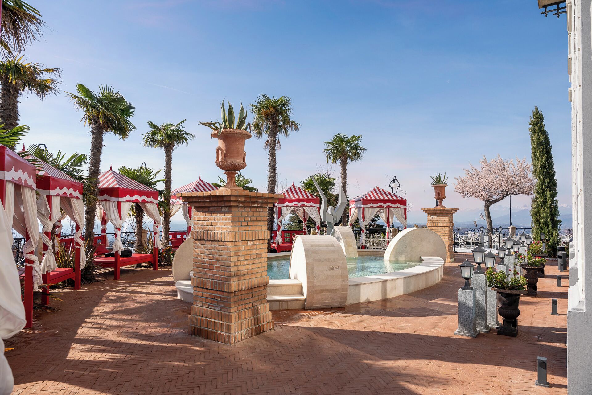 A pool area with red and white striped canopies, palm trees, and a view of the sea.