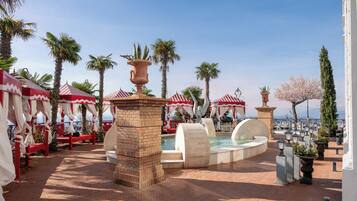 A pool area with red and white striped canopies, palm trees, and a view of the sea.