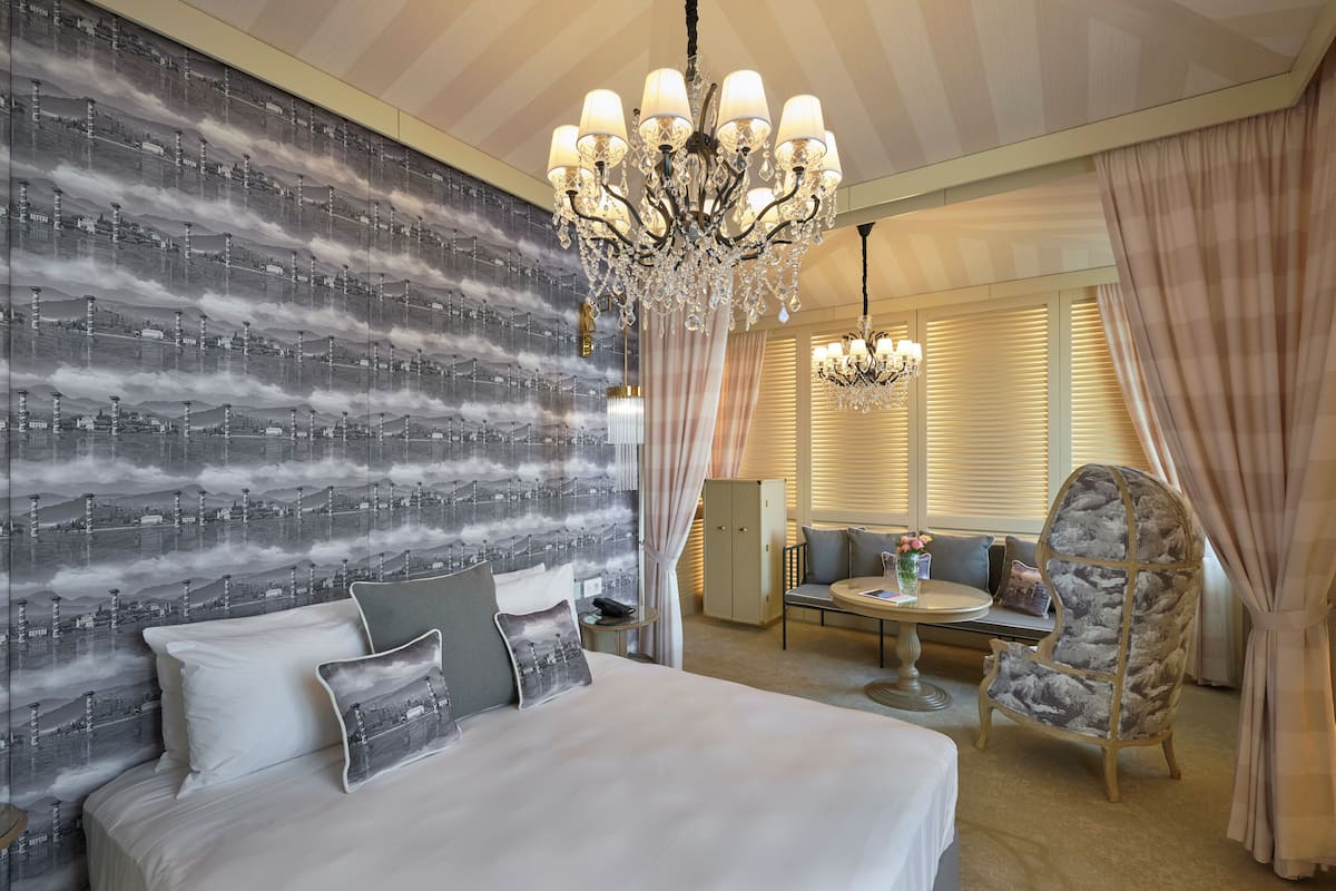 Una habitación de hotel con una cama grande, un candelabro, una zona de estar con una mesa y una silla decorativa.