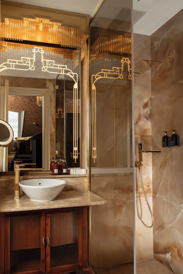 A modern bathroom with a large mirror, a white sink, and a gold faucet. There is a shower area with a glass door and a gold showerhead. - Nuvole Garden Hotel (Brunate)