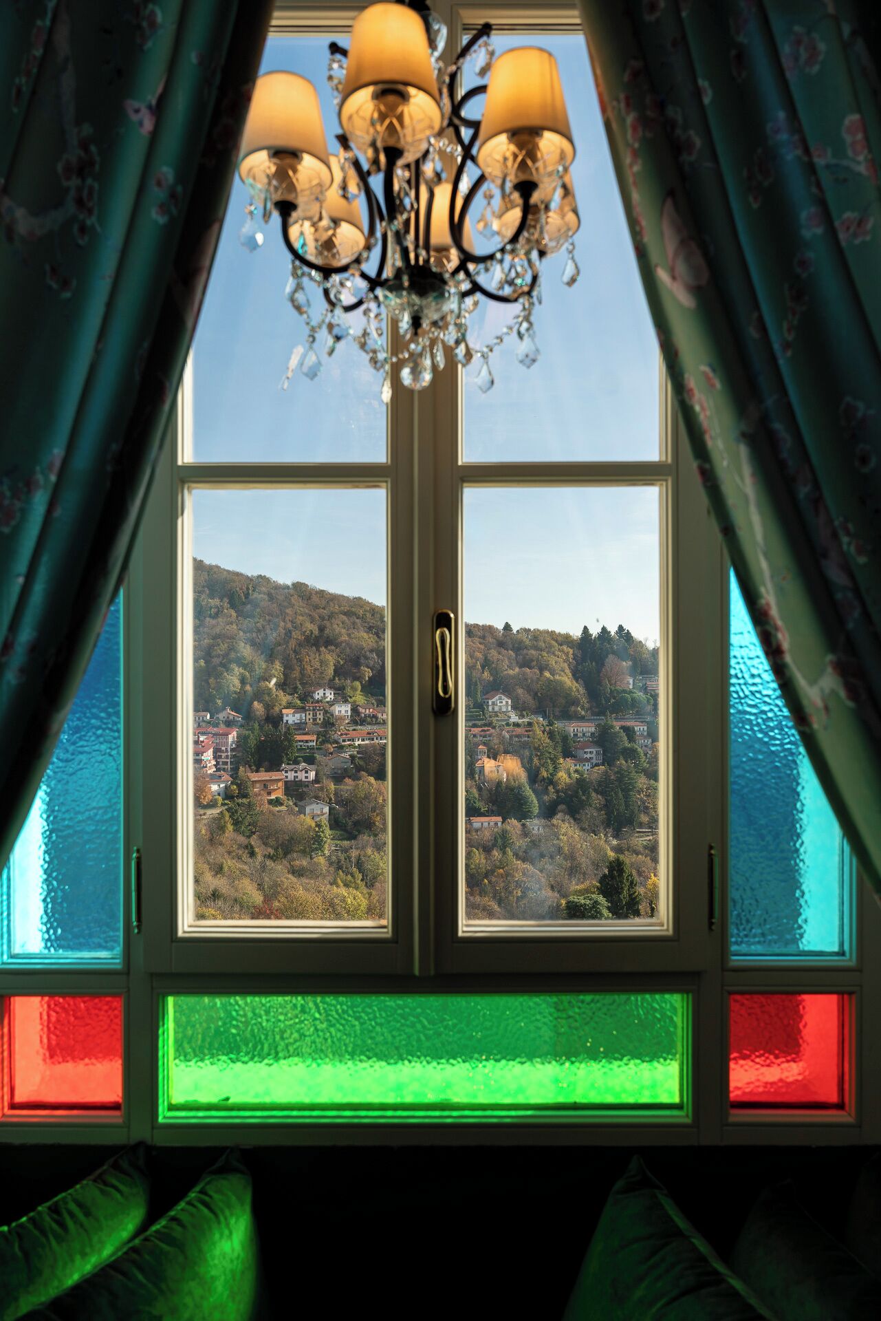 Une fenêtre donnant sur une ville perchée sur une colline, un lustre et des rideaux.