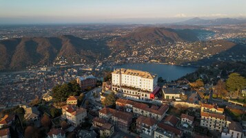 A large building with multiple floors, surrounded by residential buildings, set against a backdrop of mountains and a lake.