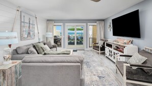 Ein modernes Wohnzimmer mit Deckenventilator, Flachbildfernseher und Blick auf den Strand durch Glastüren.