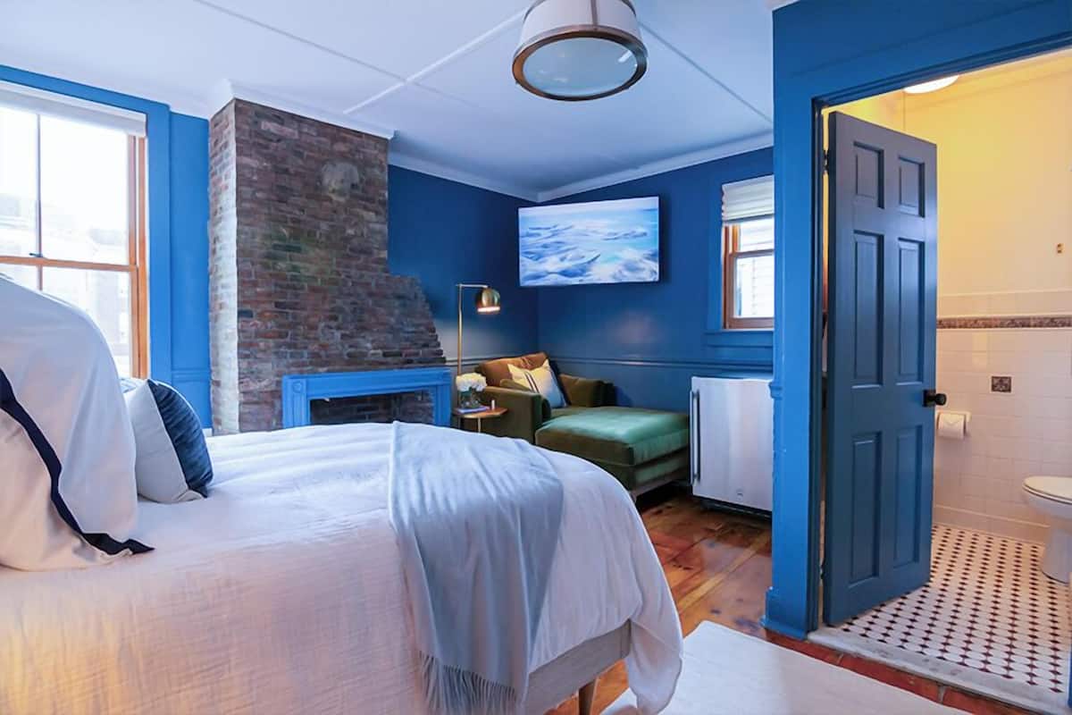 A bedroom with a bed, a brick fireplace, a television, and a door leading to a bathroom.