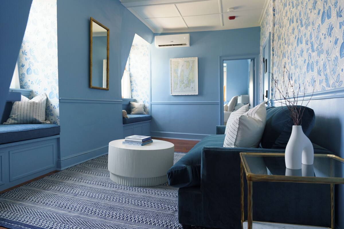 A room with a blue wall, a round white ottoman, a dark blue sofa, and a small white side table with a vase.