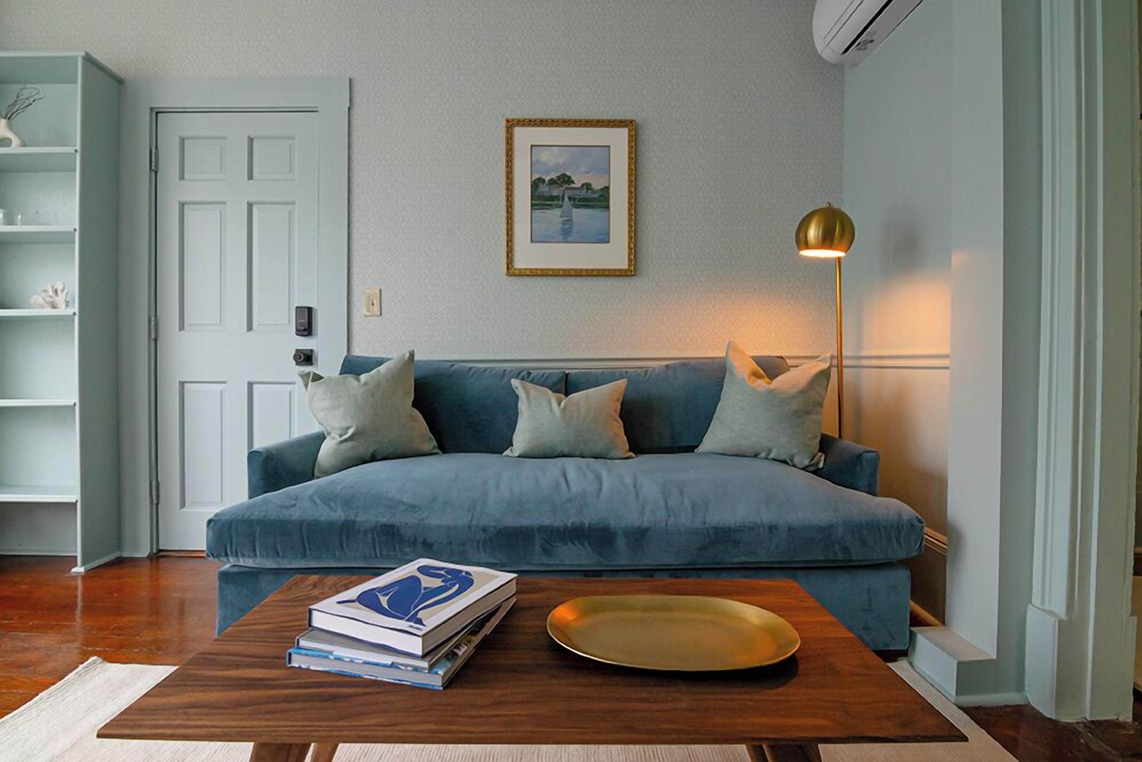 Un salon avec un canapé bleu, une table basse en bois sur laquelle sont posés des livres et un plateau, un tableau encadré au mur et un lampadaire.