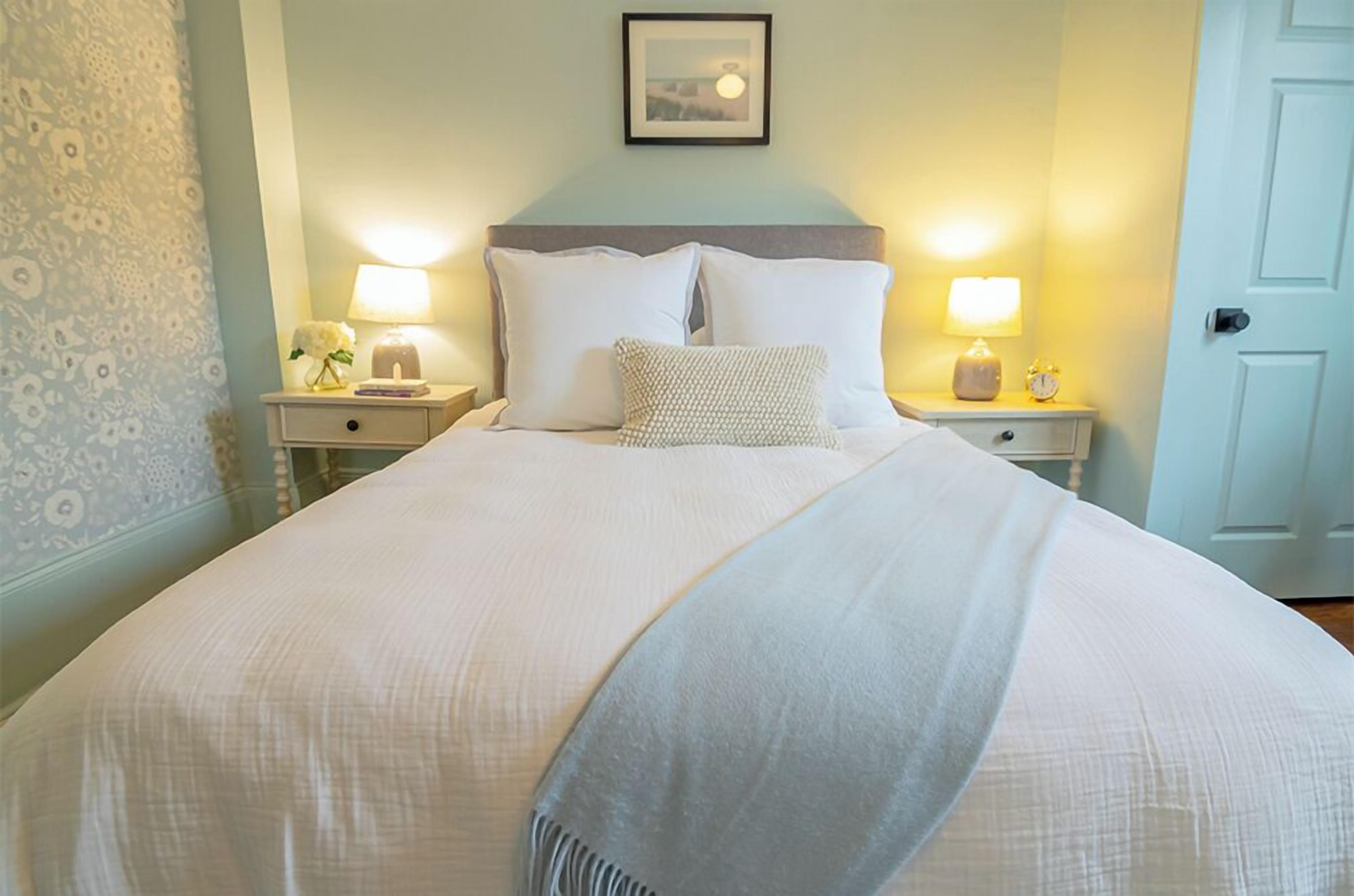 A neatly made bed with white linens and pillows, two bedside lamps, and a framed picture on the wall.