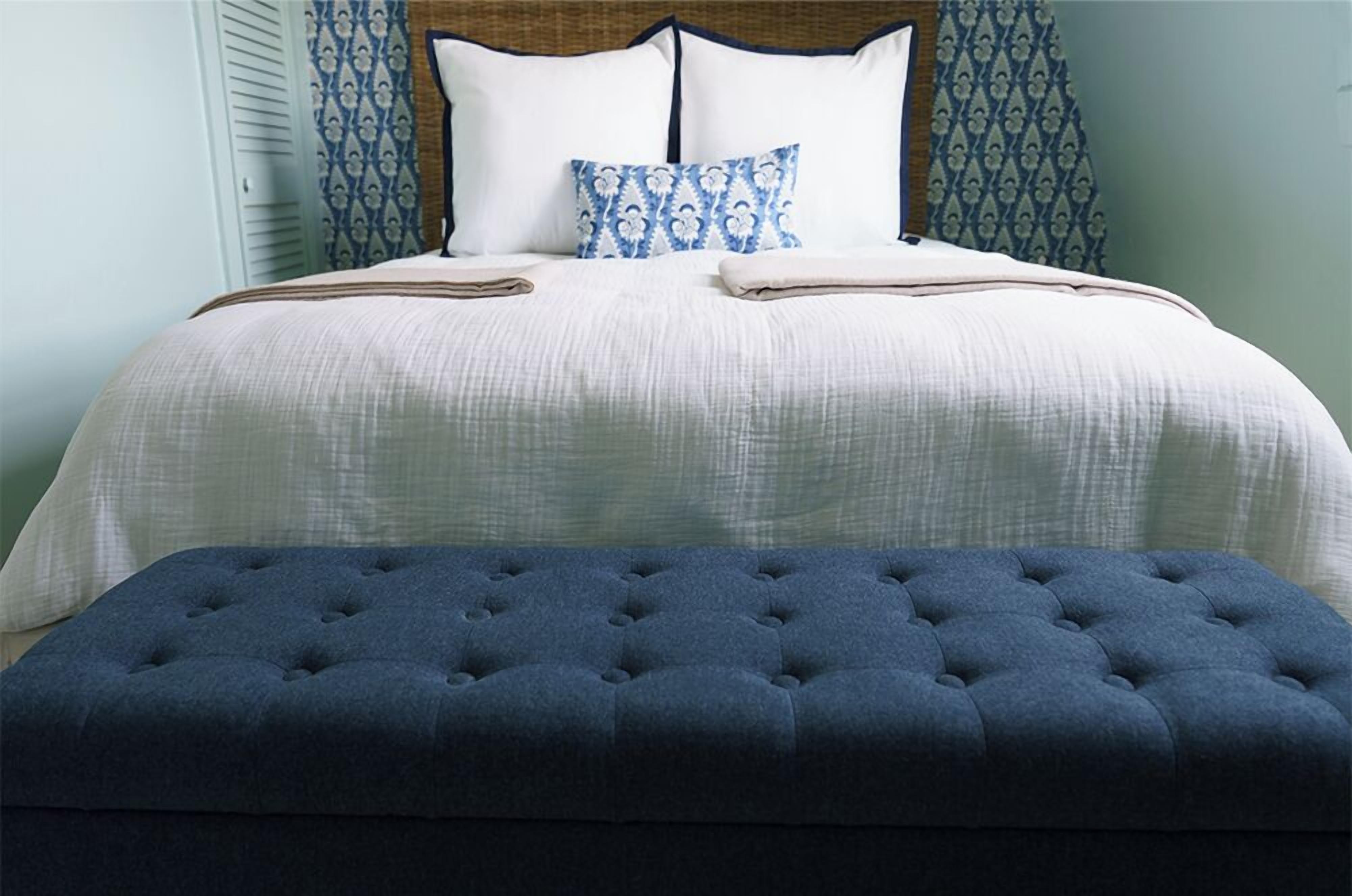 A neatly made bed with a blue upholstered bench in front, a patterned pillow, and a headboard with a wooden panel.