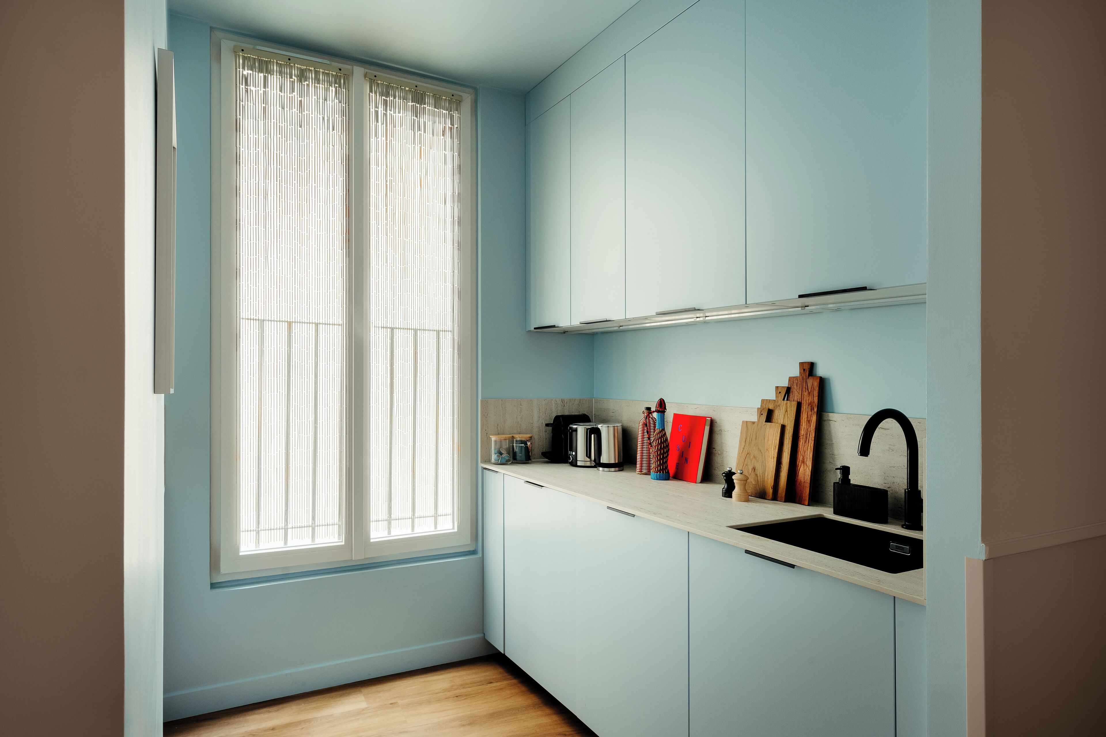 A small kitchen with light blue cabinets, a white countertop, a black sink, and a window with sheer curtains.