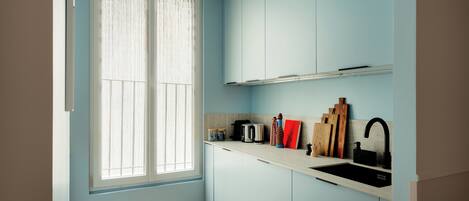 A small kitchen with light blue cabinets, a white countertop, a black sink, and a window with sheer curtains.