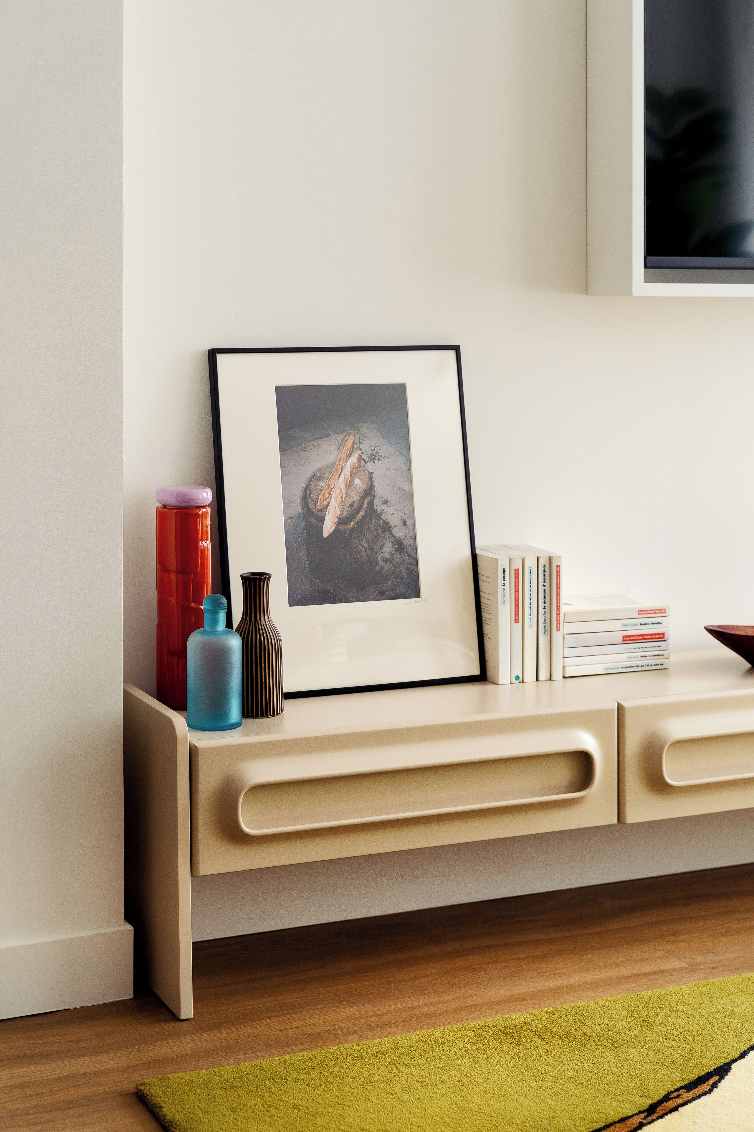 Une pièce au décor minimaliste, avec un plancher en bois, un tapis vert, une console de couleur beige, un tableau encadré, un thermos rouge, une bouteille bleue, un vase marron et une pile de livres.