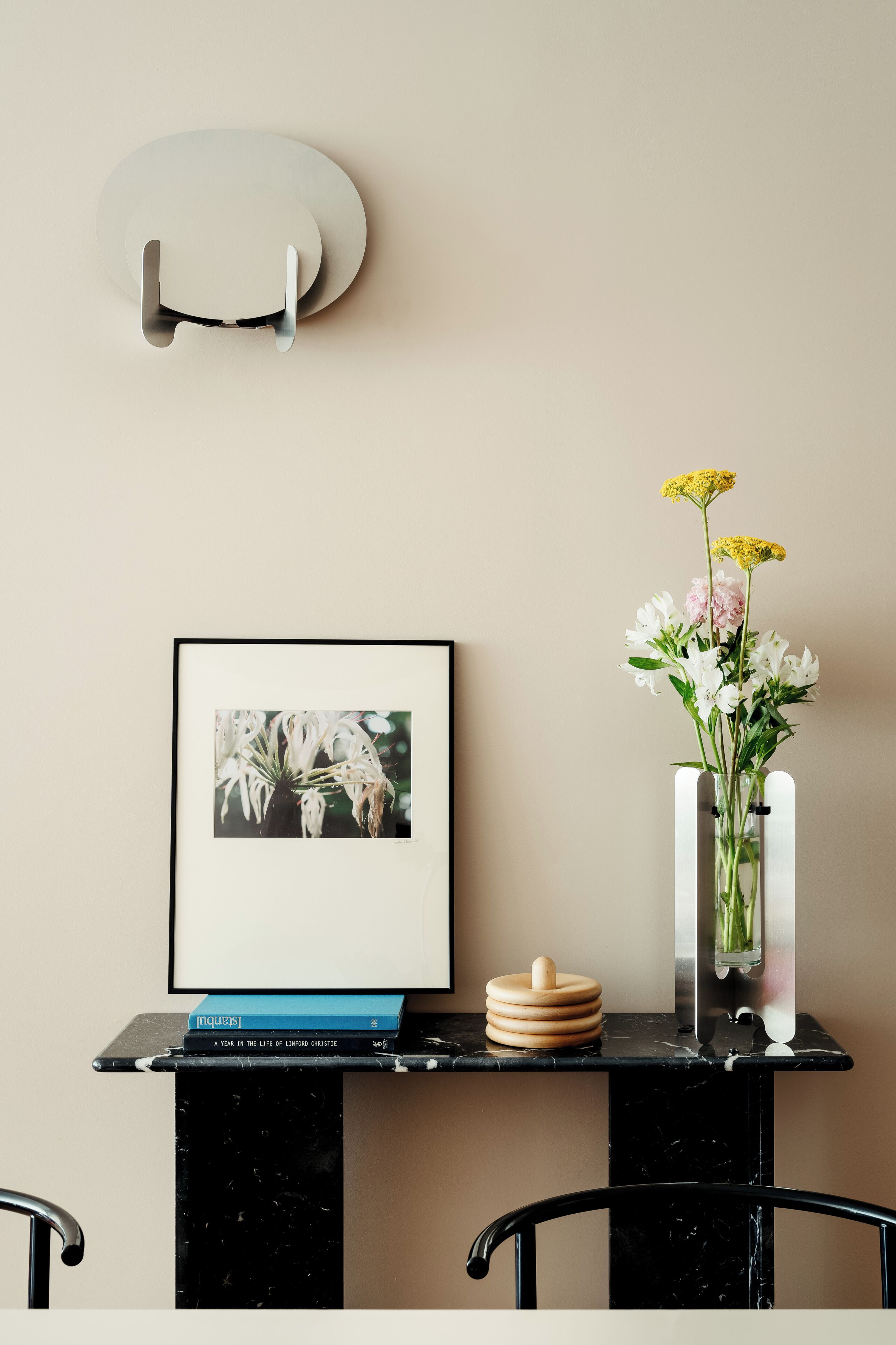 Habitación minimalista con una fotografía enmarco, una pila de libros, un florero con flores y un aplique de pared.