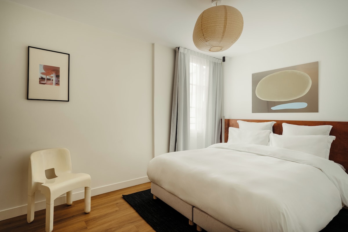 A hotel room with a bed, a chair, a picture on the wall, and a large window with curtains.