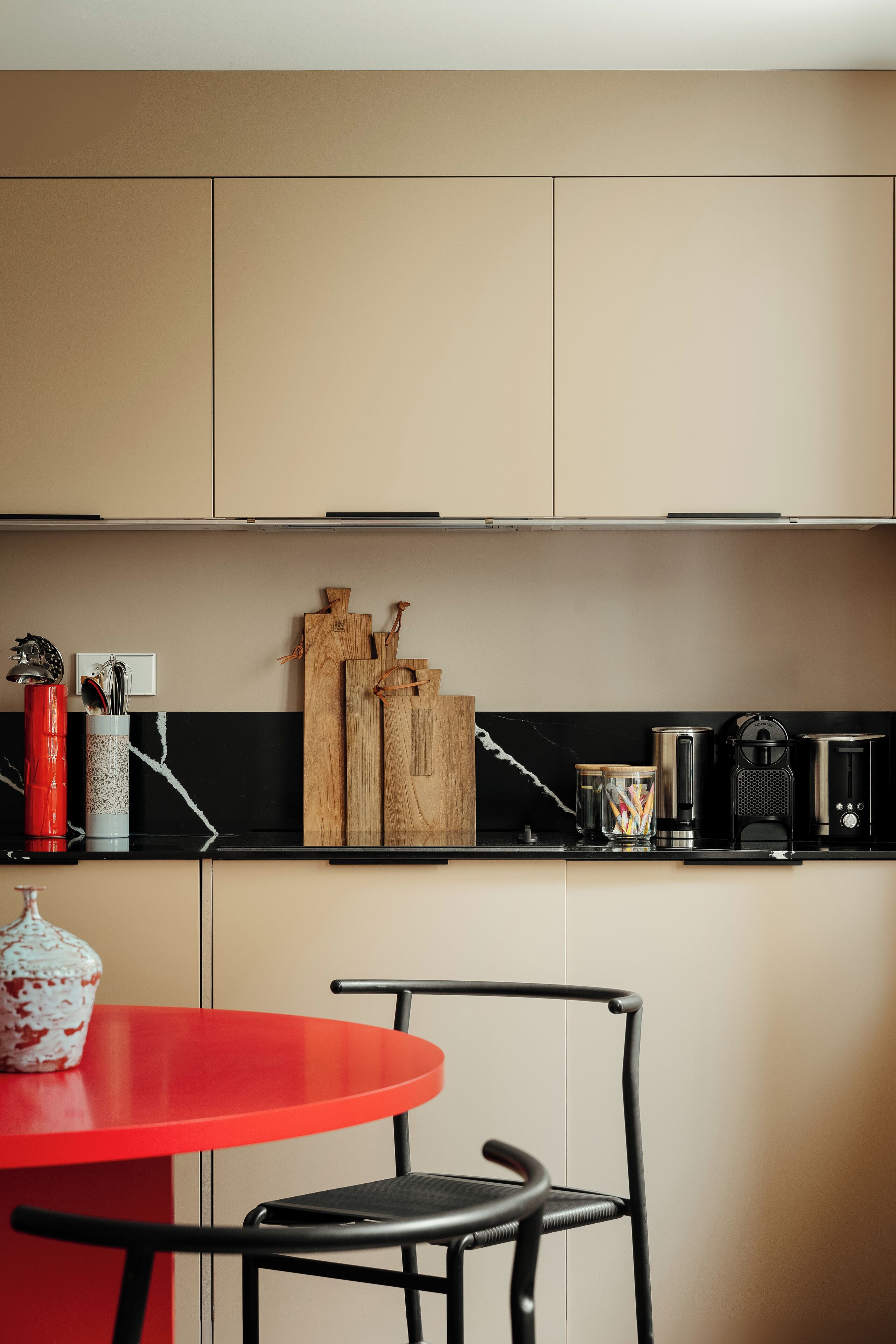 A modern kitchen with a red table and black chairs, a coffee machine, and wooden utensils on the counter.