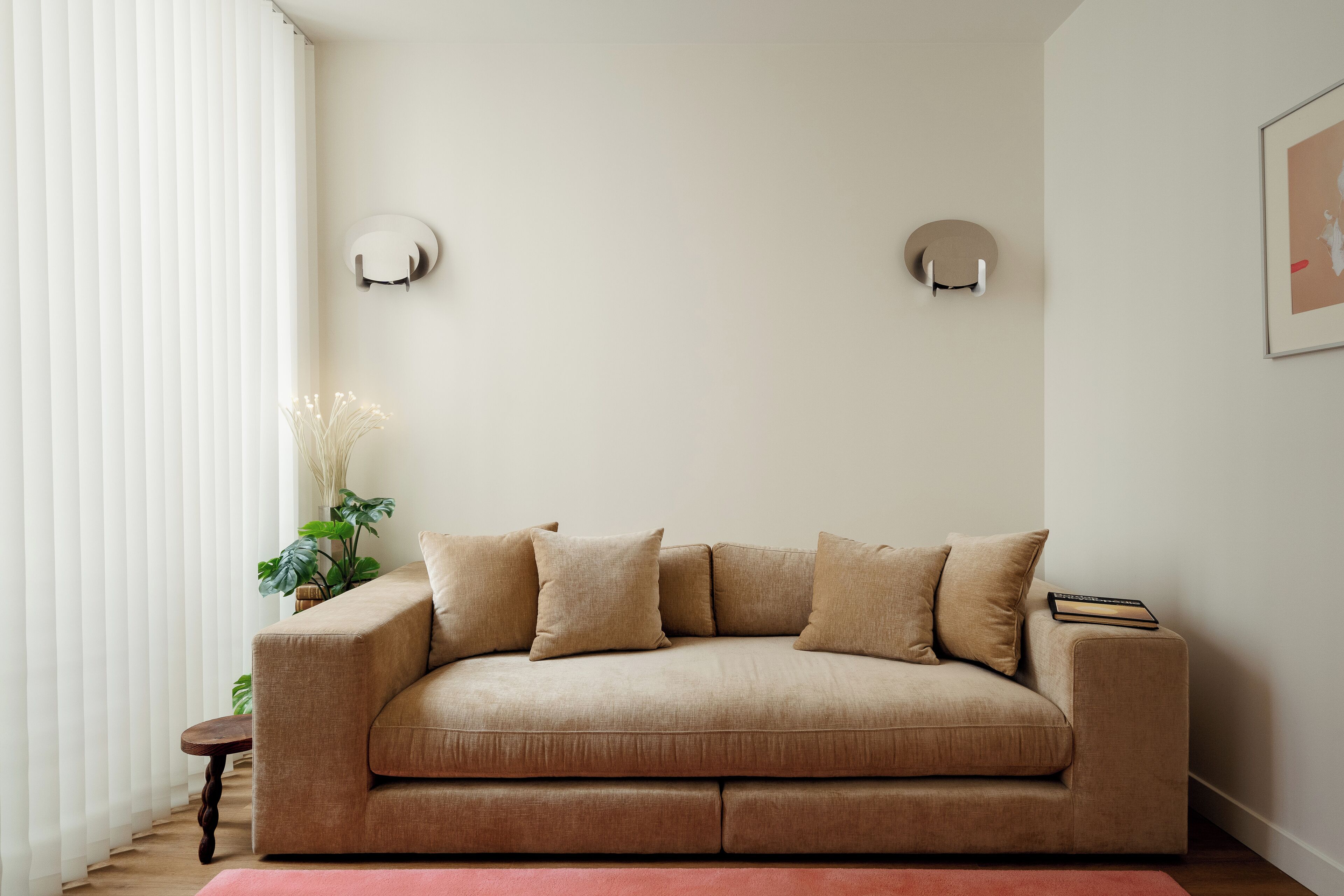 A neutral-toned living room with a beige sofa, two wall-mounted light fixtures, a small side table, and a potted plant.