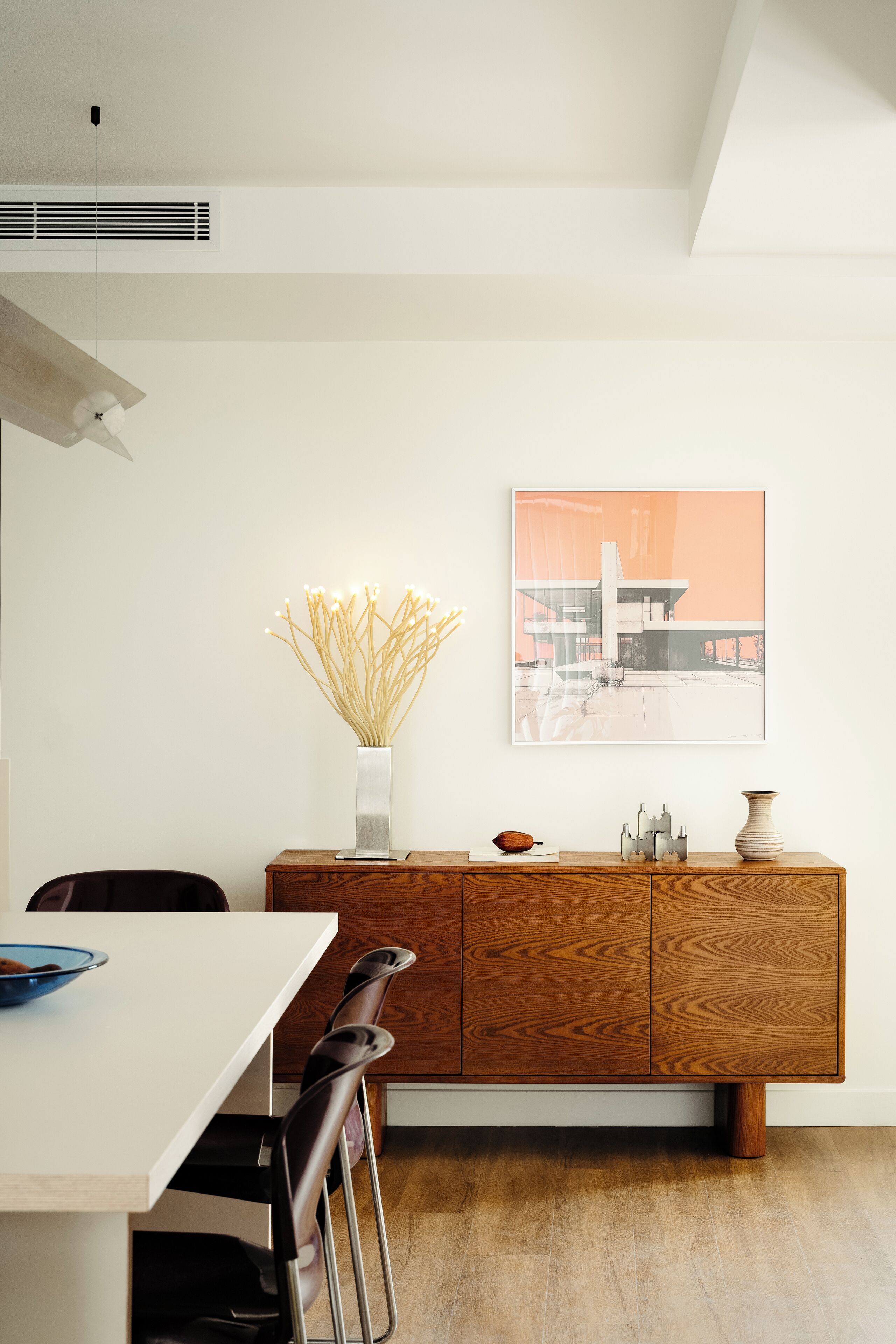 A modern kitchen with a white table, wooden cabinet, and a vase with dried plants.