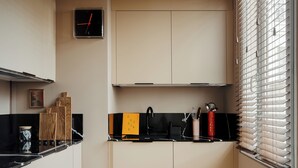 A compact kitchen with a black countertop, wooden cabinets, a clock, and a window with blinds.