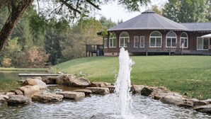 A tranquil pond with a central water feature surrounded by rocks, a gazebo in the background, and a grassy area. - Spacious Mtn Retreat on 40+ Acres (Horse Shoe)