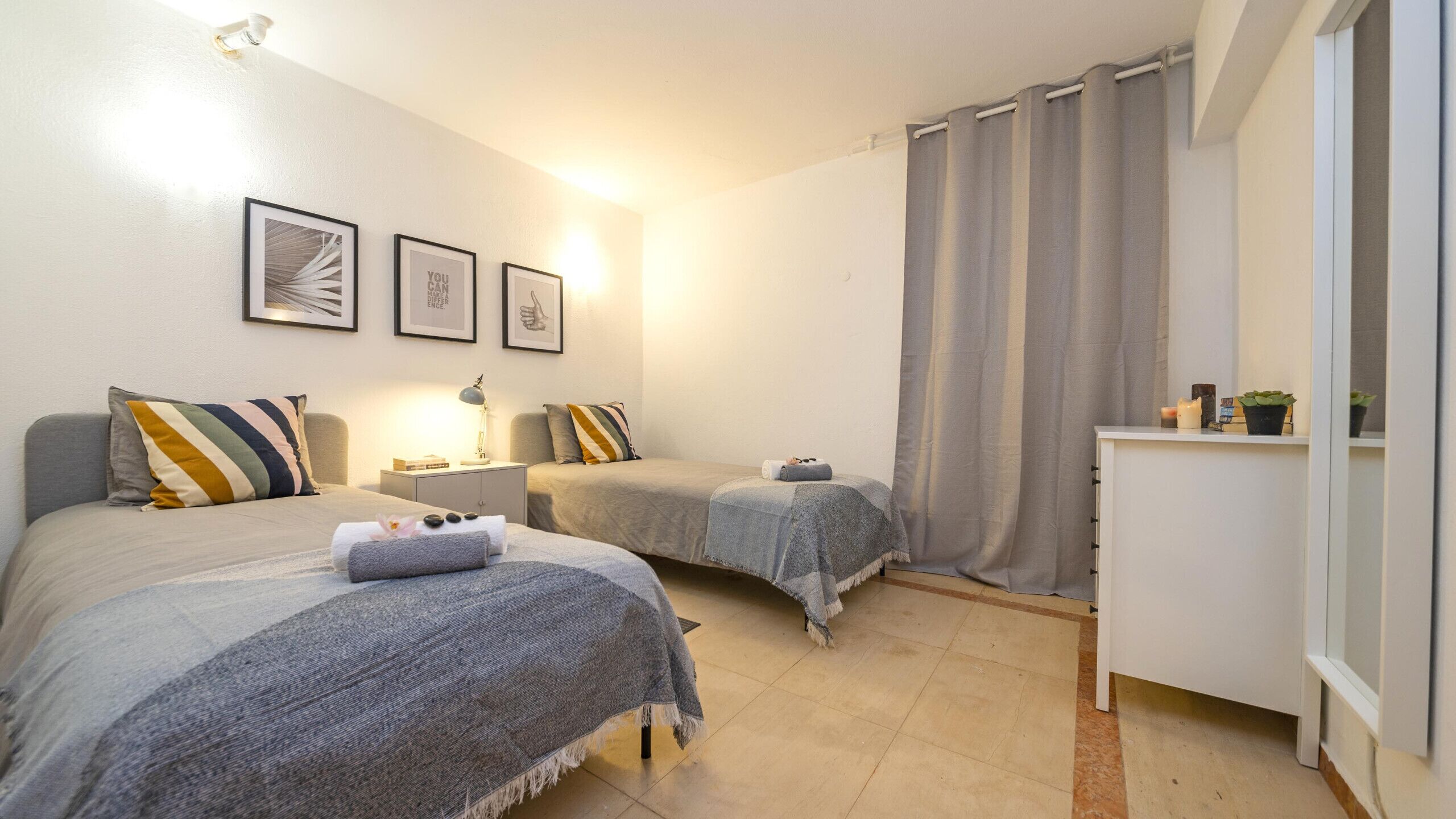 Een slaapkamer met twee bedden, een nachtkastje, een lamp, een klein kastje en een raam met gordijnen.