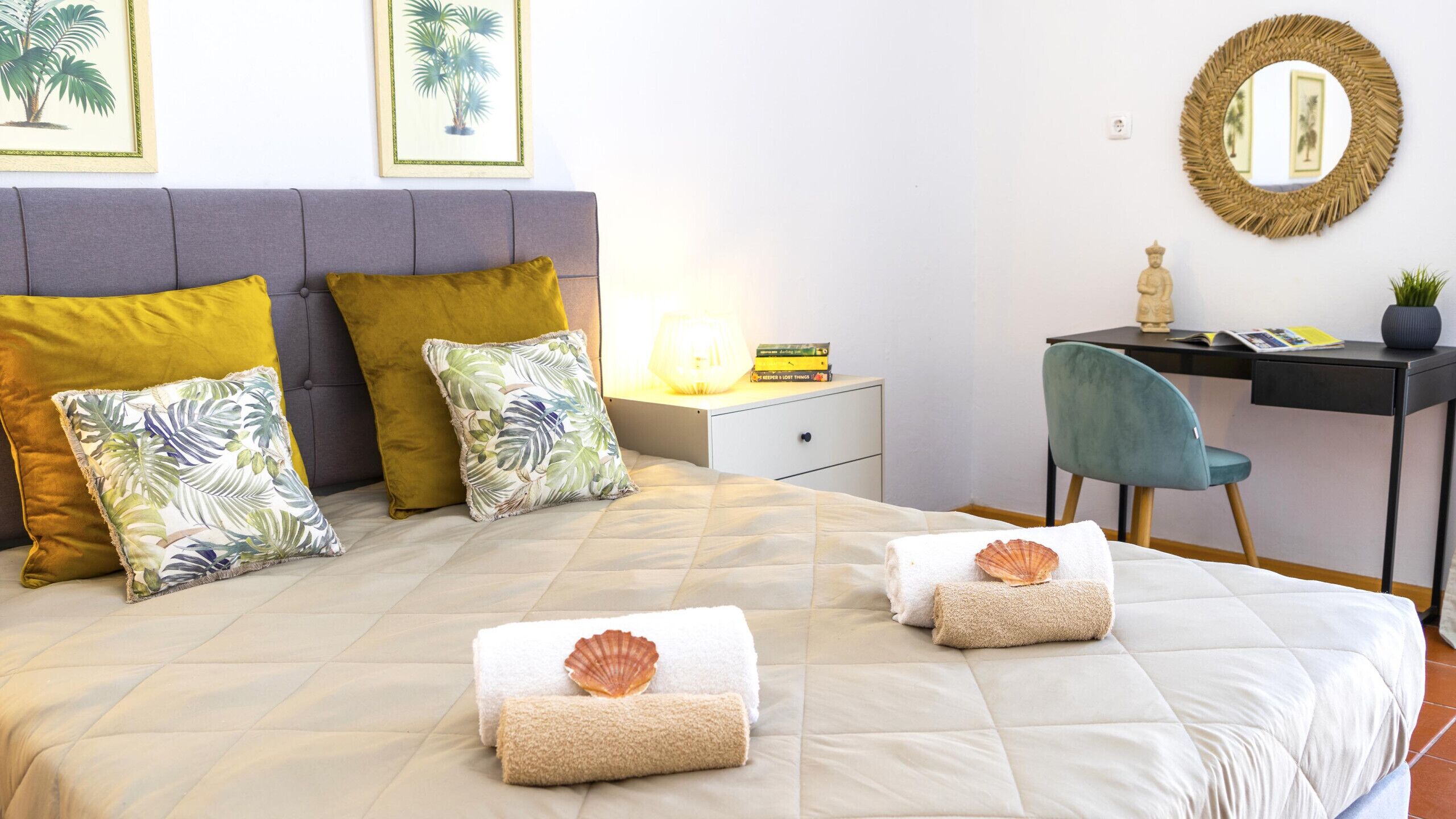 A neatly made bed with a quilted cover, two pillows with tropical leaf patterns, and two folded towels with seashells. A desk with a chair, a lamp, and a small statue are visible.