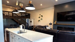 A modern kitchen with a central island, a dining area with chairs, a dark leather sofa, a staircase, and a flat-screen TV mounted on the wall. - Big Wolf Barn: Where Big Groups Have Big Fun, Lakegfront in Jackson, MI. (Jackson)