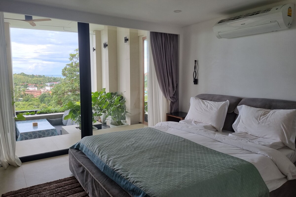 A bedroom with a large bed, a view of greenery and buildings, and an air conditioning unit on the wall.