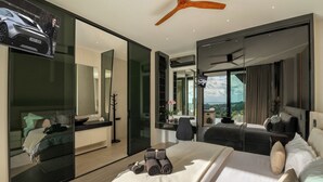 A modern hotel room with a large bed, a desk, a chair, a TV, and a view of the outdoors. - Magic Suites (Koh Samui)