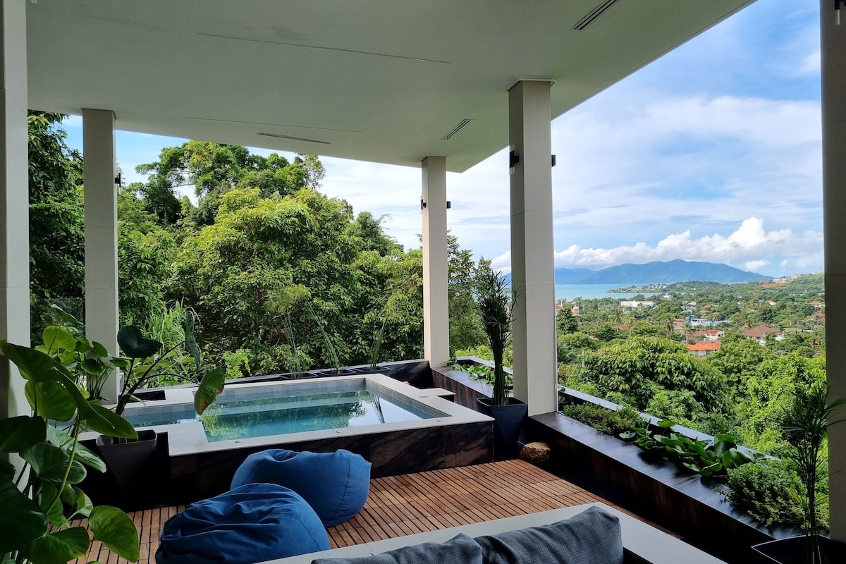 A modern outdoor lounge area with a wooden deck, blue and gray cushions, and a hot tub.