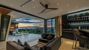 A modern living room with a sofa, a coffee table with candles, a bar area with glasses, and a view of the landscape through large windows. - Magic Suites (Koh Samui)