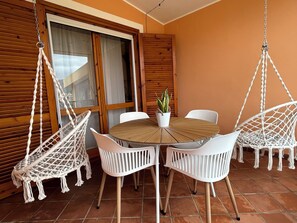 Ein runder Holztisch mit weißen Stühlen, ein hängender Makramee-Sessel und ein Blumentopf.