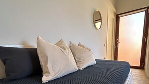 Un lit recouvert d’une couverture à motifs sombres, avec deux oreillers, une bouche d’aération fixée au mur et une porte donnant sur une autre pièce.