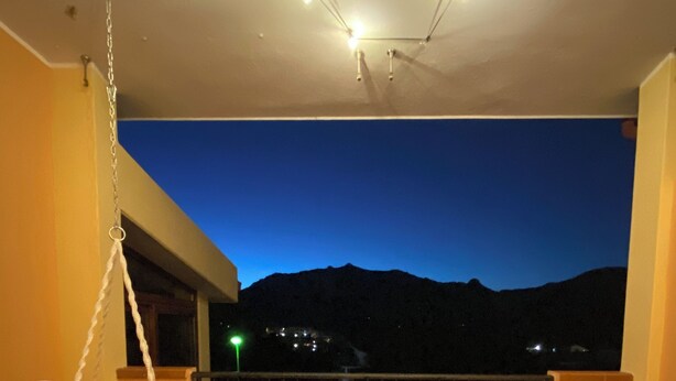 Une table ronde en bois, entourée de chaises blanches, sur un balcon offrant une vue sur les montagnes au crépuscule.