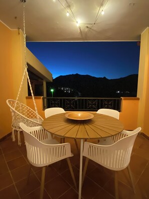 Ein runder Holztisch mit weißen Stühlen auf einem Balkon mit Blick auf die Berge in der Abenddämmerung.
