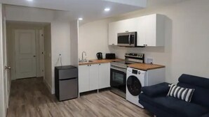 A compact kitchen with a microwave, oven, and washing machine, a small fridge, and a dark-colored sofa.