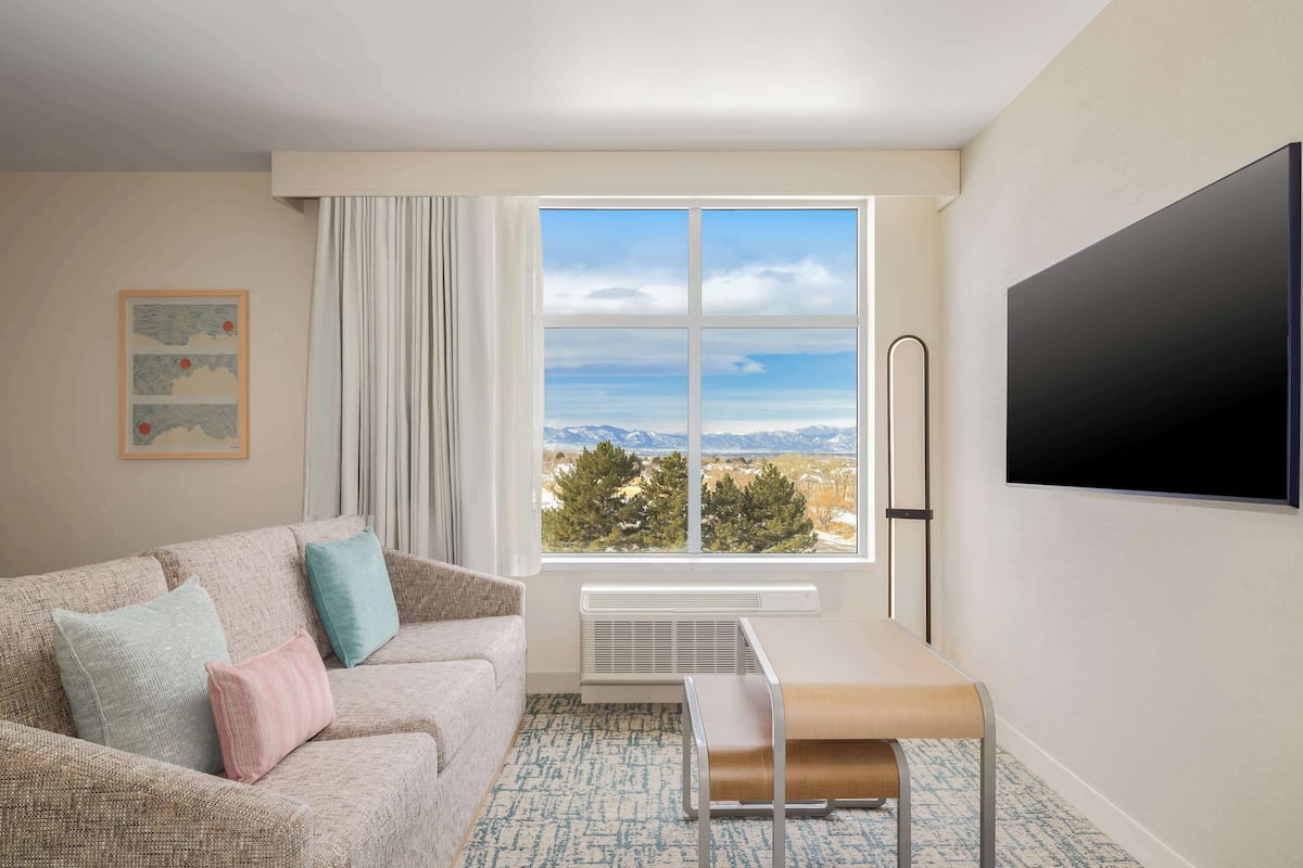 A modern living room with a sofa, a bench, a large window with a mountain view, a flat-screen TV, and a framed picture on the wall.