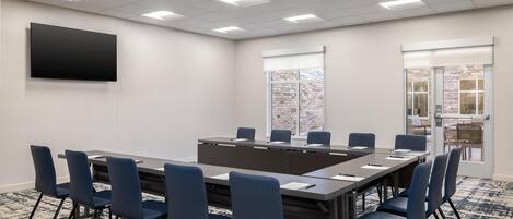 A conference room with a long table, blue chairs, a flat-screen TV, and a patterned carpet.
