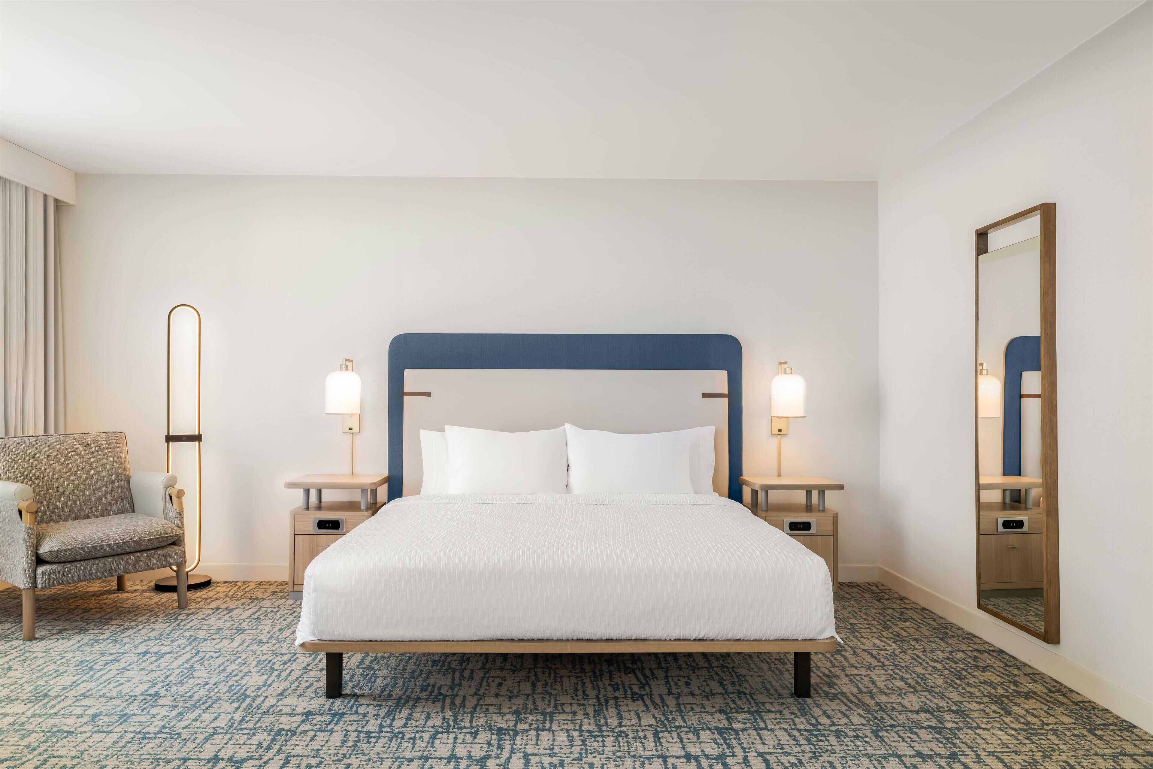 A hotel room with a large bed, two bedside tables, a chair, a floor lamp, and a mirror.