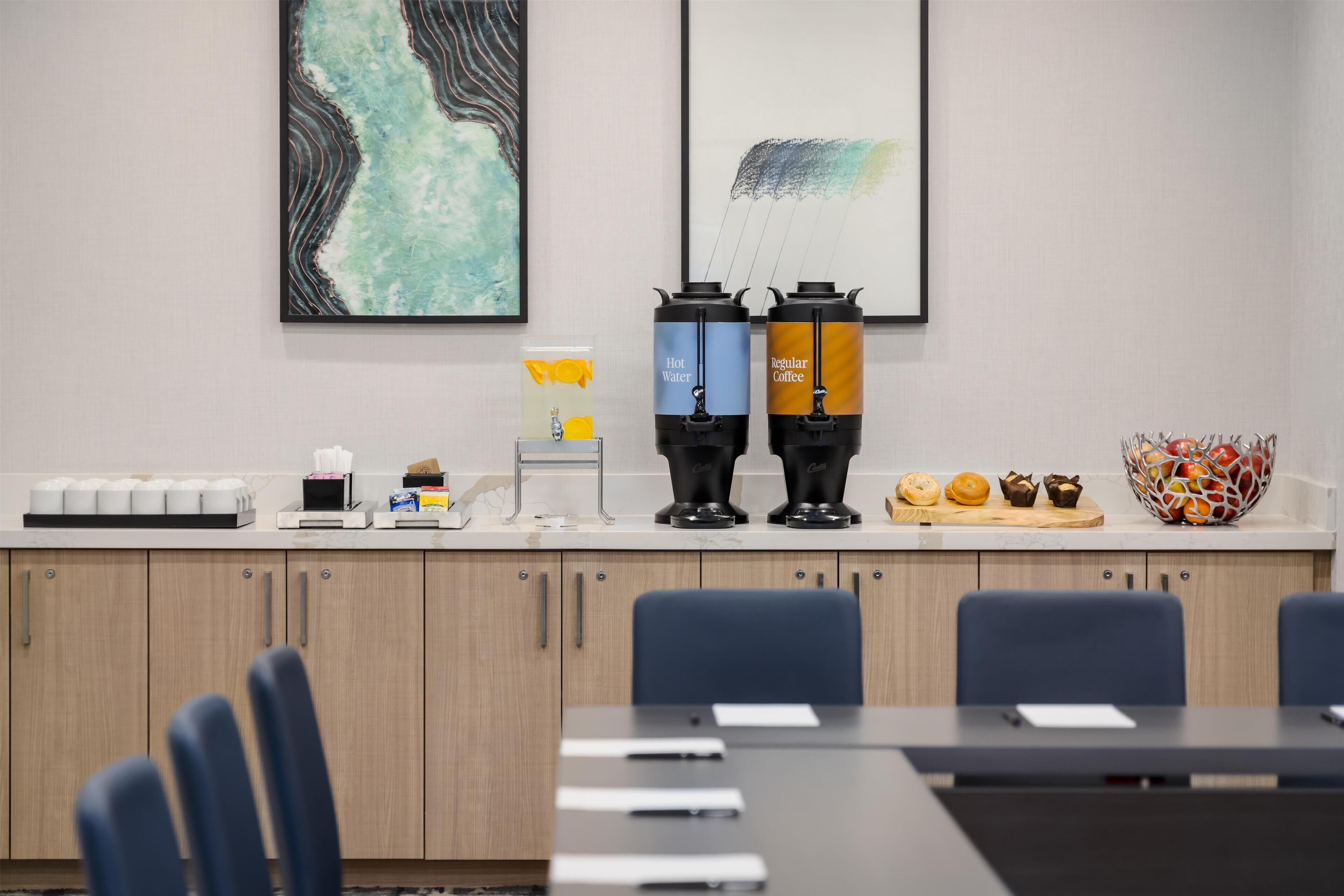 Une salle de réunion moderne équipée d’une table de conférence, de chaises et de deux distributeurs de boissons, l’un indiquant « Eau chaude » et l’autre « Café ordinaire ».