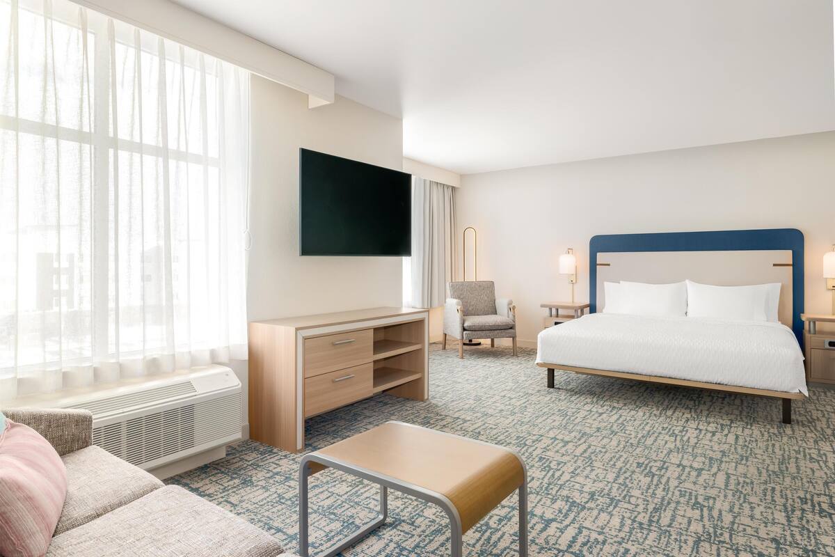 A hotel room with a bed, a TV mounted on the wall, a desk, a chair, and a window with curtains.